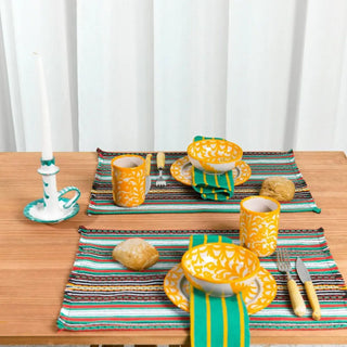 Mesa con dos manteles individual de rayas coloridas y vajilla de cerámica de granada amarilla, con tazas, platos y pan rústico, en un ambiente acogedor. Real Fábrica.