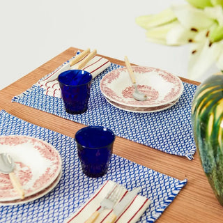 Mesa con dos manteles individual ojo de perdiz azul y vajilla de cerámica roja, con vasos azul, cubiertos y un jarrón con flores, en un ambiente acogedor. Real Fábrica.