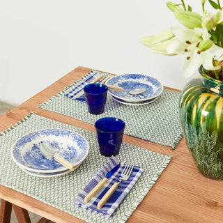 Mesa con dos manteles individual ojo de perdiz verde y vajilla de cerámica azul, con vasos, cubiertos y un jarrón con flores, en un ambiente acogedor. Real Fábrica.