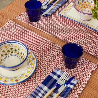 Mesa con dos manteles individual ojo de perdiz rojo y vajilla de cerámica, con vasos, cubiertos que la decoran en un ambiente acogedor. Real Fábrica.