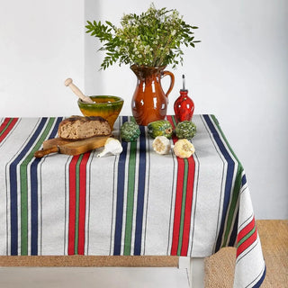 Un mantel de rayas verdes, azules y rojas crea una base fresca y alegre. Sobre la mesa, una aceitera y un mortero con ajo, junto a una tabla con pan y pimientos y un jarrón con flores, completan una escena natural y acogedora. Real Fábrica.