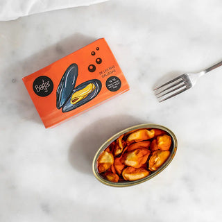 Foto cenital muestra un mejillón con su caja de presentación y delante un mejillón abierto con un tenedor a su lado, es de las  las Rías Gallegas. Real Fábrica.