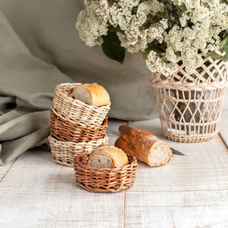 Paneras redondas de mimbre tostado y blanco, ideales para la decoración de mesas y servicio de pan, a su lado lo adorna unas flores blancas en un jarrón de mimbre. Real Fábrica.