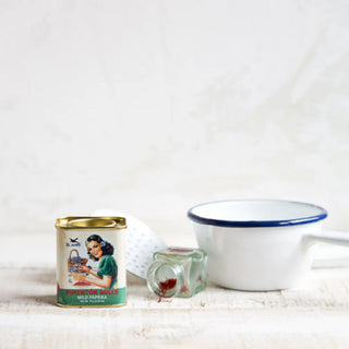 Foto frontal de una lata de pimentón dulce, junto a un pequeño frasco de vidrio volcado con hebras de especia y un cuenco de peltre blanco, sobre una superficie clara. Real Fábrica.