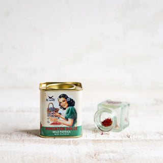 Foto frontal de lata de pimentón dulce, junto a un pequeño frasco de vidrio volcado con hebras de especia sobre una superficie clara. Real Fábrica.