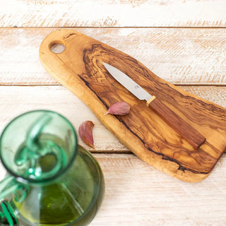 Foto cenital de una tabla de cortar de madera con un cuchillo de cocina y dos dientes de ajo sobre una mesa rústica blanca; en primer plano aparece una aceitera de vidrio verde con aceite de oliva. Real Fábrica.
