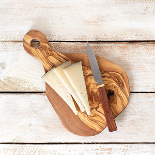 Foto cenital de una tabla de cortar de madera con un cuchillo de cocina y tres trozos de queso. Real Fábrica.