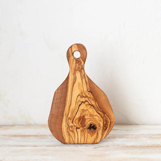 Foto frontal de tabla mini redonda de madera de olivo en posición vertical sobre una mesa rústica blanca. Real Fábrica.