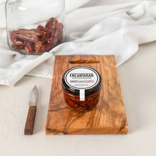 Foto de un tarro de cristal de tomates secos sobre una tabla de madera y a su lado un cuchillo. Real Fábrica.