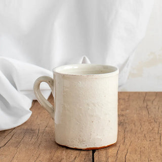 Foto de un vaso de cerámica artesanal con asa y acabado de esmalte blanco craquelado, sobre una mesa de madera y de fondo una cortina blanca. Real Fábrica.
