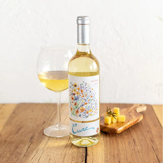 Fotografía de un vino blanco afrutado de Cune, con una copa al lado y una tabla de madera con queso al fondo. Real Fábrica.