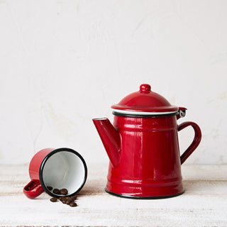 Cafetera pequeña de peltre esmaltado roja junto a una taza esmaltada con granos de café.