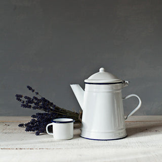 Taza blanca mini de peltre junto a una cafetera esmaltada blanca con filo azul y un ramo de lavanda.