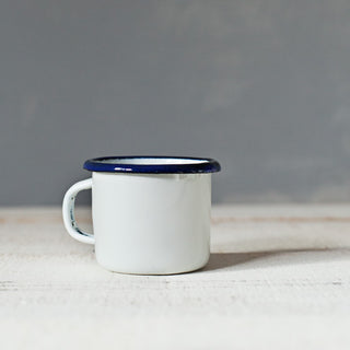 Taza blanca mini de peltre esmaltado con filo azul sobre superficie de madera.
